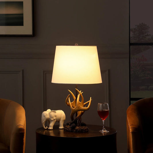 Natural antlers lamp with white shade on table between two brown chairs with elephant figurine and wine glass